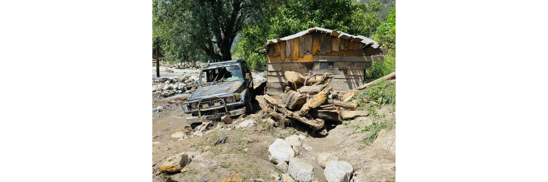 Un véhicule couvert de boue à côté d’une structure en bois endommagée, entourée de débris, de rochers et de troncs d’arbres tombés après des inondations en Afghanistan.