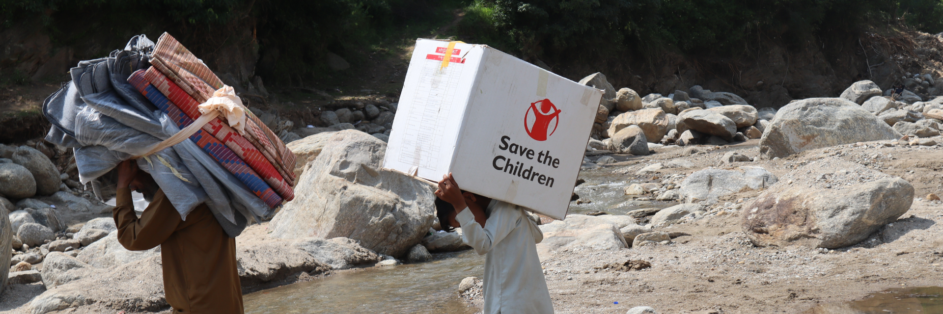 Deux hommes transportent des provisions à travers un ruisseau. L'un porte des textiles, l'autre une boîte sur laquelle est inscrit « Save the Children ».