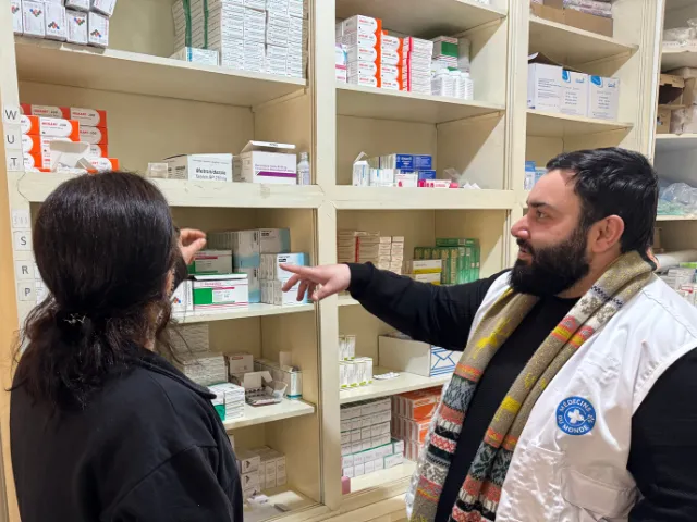 Man wearing a Médecins du Monde vest gestures at shelves of medical supplies while talking to a woman.