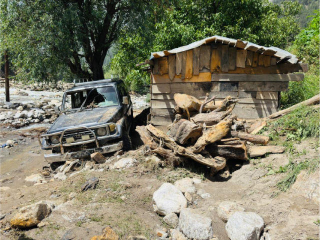 Un véhicule couvert de boue à côté d’une structure en bois endommagée, entourée de débris, de rochers et de troncs d’arbres tombés après des inondations en Afghanistan.