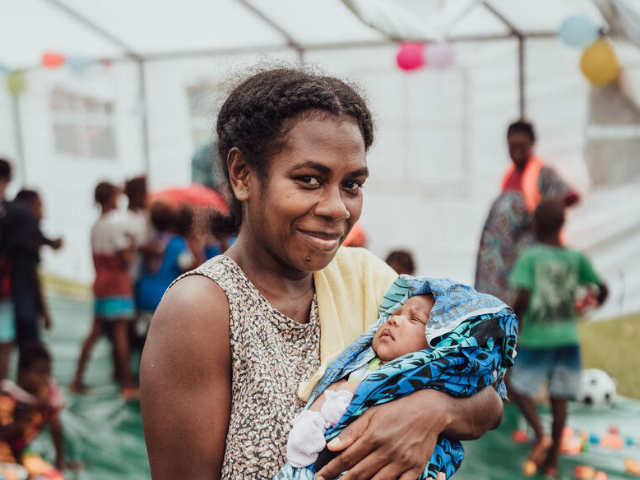 Rael et son bébé se tiennent devant une tente où des enfants jouent.