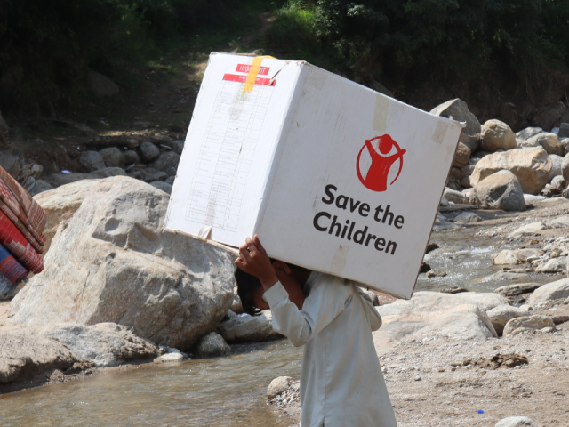 Deux hommes transportent des provisions à travers un ruisseau. L'un porte des textiles, l'autre une boîte sur laquelle est inscrit « Save the Children ».